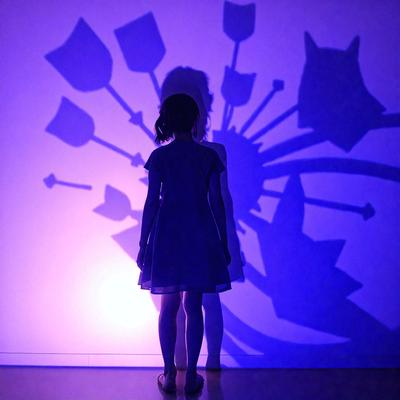 The primary subject of the image is a young girl standing in front of a wall with a shadow of a flower pattern on it. The setting appears to be a room with a purple light, creating a unique and artistic atmosphere. The visual style of the image is a combination of a photo and a digital art, as it captures the girl's shadow and the flower pattern on the wall. The colors and mood of the image are predominantly purple, which adds to the overall artistic and creative vibe of the